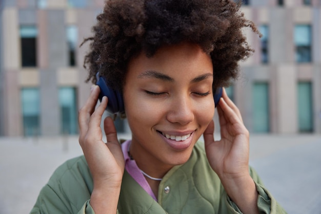 Close-up shot van een vrolijke vrouw met krullend haar sluit de ogen van plezier terwijl het favoriete audionummer via een koptelefoon een jas draagt