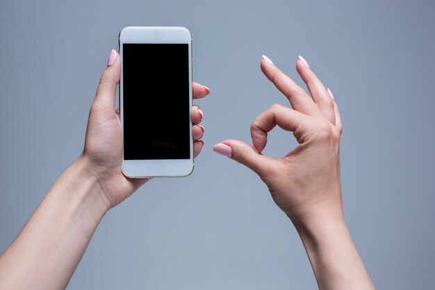 Close-up shot van een vrouw te typen op de mobiele telefoon op een grijze achtergrond. Vrouwelijke handen met een moderne smartphone en wijzen met figer. Leeg scherm om het op uw eigen webpagina of bericht te plaatsen.