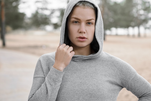 Close-up shot van fit zelfverzekerde jonge vrouw die actieve gezonde levensstijl kiest, buitenshuis traint om een perfecte lichaamsvorm te krijgen en af te vallen, poseren geïsoleerd in stijlvolle hoodie, camera kijken