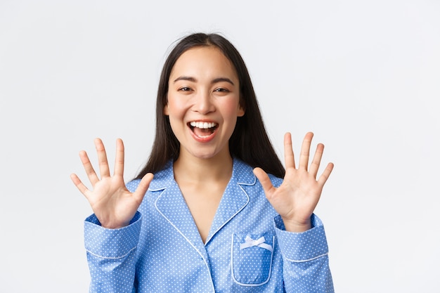 Close-up shot van gelukkige aantrekkelijke aziatische vrouw in blauwe pyjama met tien vingers en lachende witte tanden, uitleg van de belangrijkste regels of orde maken, staande witte achtergrond, product aanbevelen