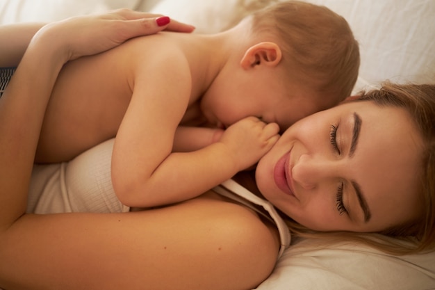 Close-up shot van gelukkige charmante jonge moeder liggend op bed met haar slaperig kindje op de borst. Mooie moeder en baby knuffelen in de slaapkamer. Liefde, geluk, moederschap en ouderschap concept