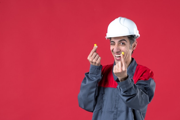 Close-up shot van gelukkige jonge bouwer in uniform met harde hoed en met oordopjes op geïsoleerde rode muur