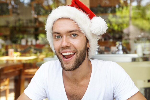 Close-up shot van gelukkige jonge hipster met stijlvolle baard dragen witte t-shirt en rode kerstman hoed, Kerstmis vieren in warme tropische land, ontspannen in café tegen onscherpe achtergrond