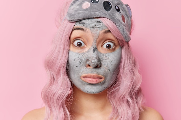Close-up shot van geschokte vrouw draagt een voedend moddermasker voor verjonging ondergaat dagelijkse schoonheidsroutines staart afgeluisterde ogen heeft lang geverfd haar geïsoleerd over roze achtergrond. Gezichtsverzorging concept