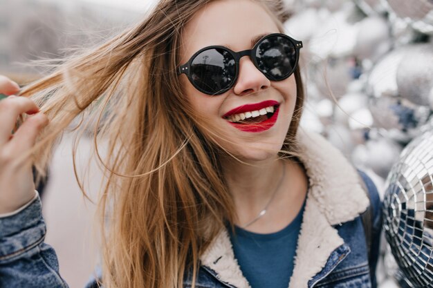 Close-up shot van innemende meisje met rode lippen spelen met haar haar en lachen op stedelijke straat. Foto van vrij Kaukasische vrouw die in zwarte zonnebril de lentegang geniet.