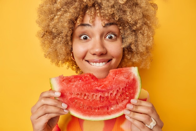 Close-up shot van krullend haired positieve vrouw houdt grote plak van sappige watermeloen geniet van het eten van haar favoriete zomerfruit bijt lippen kijkt verwonderd geïsoleerd over gele achtergrond Mmm heerlijk