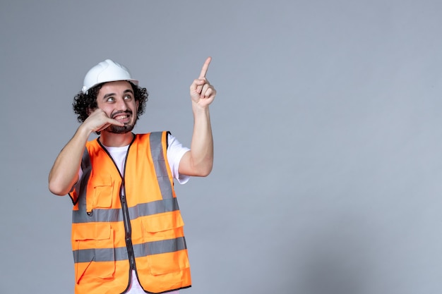 Close-up shot van lachende mannelijke constructeur in waarschuwingsvest met veiligheidshelm en omhoog wijzend aan de linkerkant en bel me gebaar over grijze golfmuur