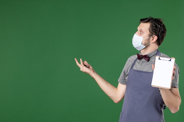 Close-up shot van nieuwsgierige mannelijke ober in uniform met medisch masker en met orderboekpen op groene achtergrond