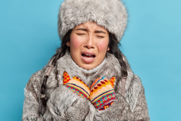 Close-up shot van ontevreden jonge aziatische inuit vrouw draagt gebreide wanten en winter outfit voelt koud aan tijdens de ijzige winter geïsoleerd over blauwe muur