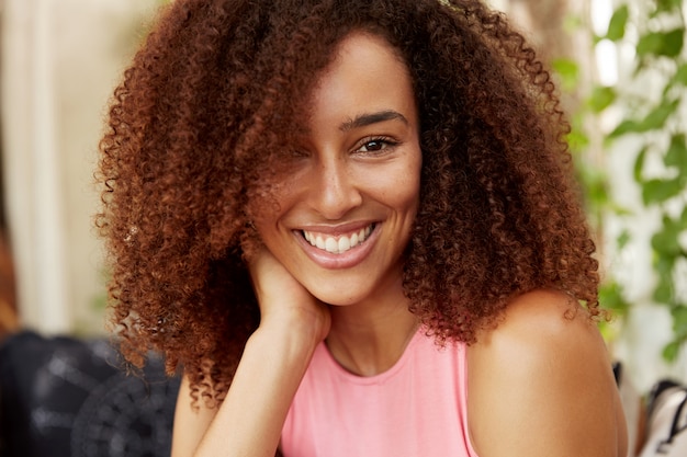 Close-up shot van positieve donkere huid tienermeisje heeft Afro kapsel, terloops gekleed, heeft een stralende glimlach, berust binnen met goede vriend of vriend, in goed humeur. Mensen, schoonheid, ethiniciteit