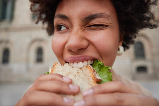 Close-up shot van positieve krullend haired blij jonge vrouw knipoogt oog houdt smakelijke sandwich met handen bijt heerlijke snack komt terug van fast food restaurnat poses buitenshuis. cheat maaltijd concept.