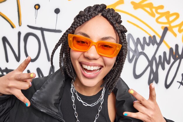 Close-up shot van positieve vrouw glimlacht toothily voelt koele gebaren actief draagt oranje zonnebril metalen kettingen om nek zwarte jas vormt tegen graffiti muur