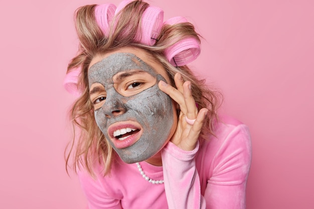Close-up shot van serieuze Europese vrouw kijkt aandachtig naar camera raakt gezicht past klei voedend masker toe voor verjonging maakt kapsel met haarrollers geïsoleerd over roze achtergrond