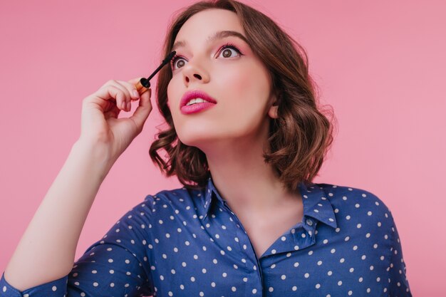 Close-up shot van spectaculaire vrouw met mascara voor wimpers en opzoeken. Binnenfoto van vrij vrouwelijk model poseren op roze muur terwijl het doen van oogmake-up vóór datum.