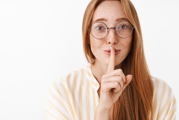 Close-up shot van tedere en vrouwelijke schattige roodharige vrouw in glazen met sproeten shh teken tonen shush geluid met wijsvinger over mond mysterieus glimlachend met liefde en zorg