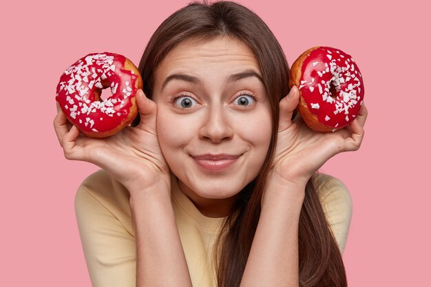 Close-up shot van tevreden mooie vrouw heeft donker haar, houdt twee rode donuts met hagelslag