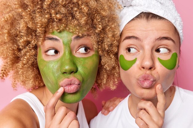Close-up shot van twee jonge vrouwen houden de lippen gevouwen, kijken naar elkaar, breng een groen voedend masker aan op het gezicht en hydrogelpleisters onder de ogen ondergaan schoonheidsprocedures poseren binnenshuis Gezichtsverzorgingsconcept