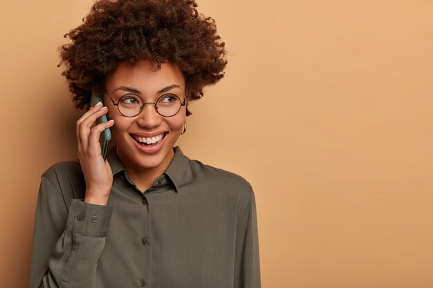 Close-up shot van vrolijke Afro-Amerikaanse vrouw heeft een hilarisch telefoongesprek, glimlacht breed en kijkt ergens peinzend