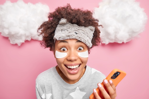 Close-up shot van vrolijke Afro-Amerikaanse vrouw in slaapmasker en pyjama kijkt graag naar camera maakt gebruik van mobiele telefoon creats post voor haar blog draagt collageen patches onder ogen vormt over wolken