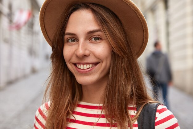 Close-up shot van vrolijke vrouwelijke avonturier draagt hoofddeksels, gestreepte trui, heeft blije uitdrukking, wandelt buiten in de promotieruimte van de stad