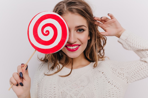 Close-up shot van vrouwelijk model met rode lippenstift met lolly op witte muur