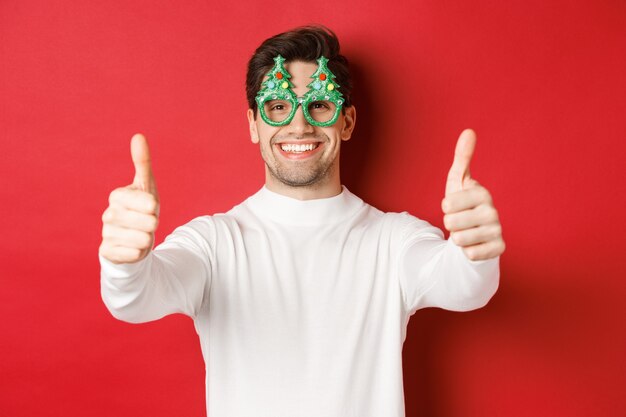 Close-up van aantrekkelijke gelukkige man in feestbril en witte trui, duimen omhoog in goedkeuring en glimlachend, staande over rode achtergrond.