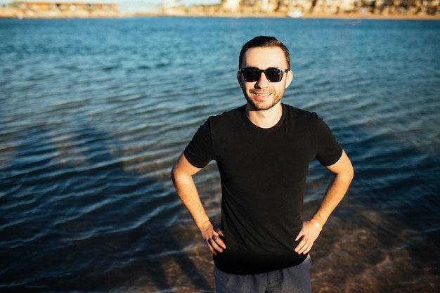 Close-up van aantrekkelijke jonge man in zonnebril in zwart t-shirt staande op het strand