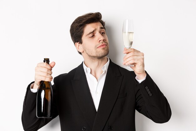 Close-up van aantrekkelijke man in trendy pak, champagne proeven, kijken naar glas, staande tegen een witte achtergrond.
