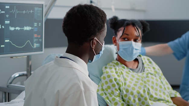 Close up van arts praten met jonge patiënt in bed, gezichtsmaskers dragen. Man aan het werk als specialist die gezondheidszorg en medicijnen bespreekt met zieke vrouw tijdens de pandemie van het coronavirus