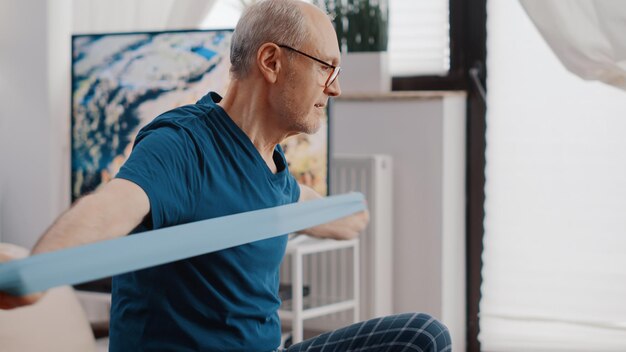 Close up van bejaarde persoon die weerstandsband trekt om te oefenen en op fitness toning bal zit. Senior man traint met elastische riem en kijkt naar video van trainingsles op laptop