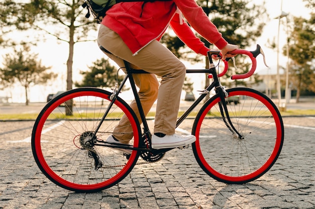 Close-up van benen in sneakers en handen op stuurwiel van hipster stijl bebaarde man in rode hoodie en beige broek alleen rijden met rugzak op fiets gezonde actieve levensstijl reiziger backpacker