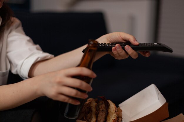 Close-up van blanke vrouw handen veranderende tv-stations met behulp van zwarte tv-afstandsbediening in de woonkamer. Persoon met bierfles ontspannen met afhaalmaaltijden hamburger voor tafel met levering fastfood.