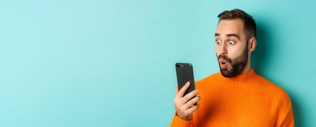 Close-up van de blanke man die naar het telefoonscherm staart met een verrast gezicht met een oranje trui licht bl