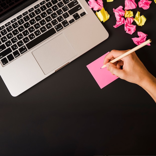 Close-up van de hand die van de vrouw op zelfklevende nota met laptop en verfrommeld document op zwarte achtergrond schrijft