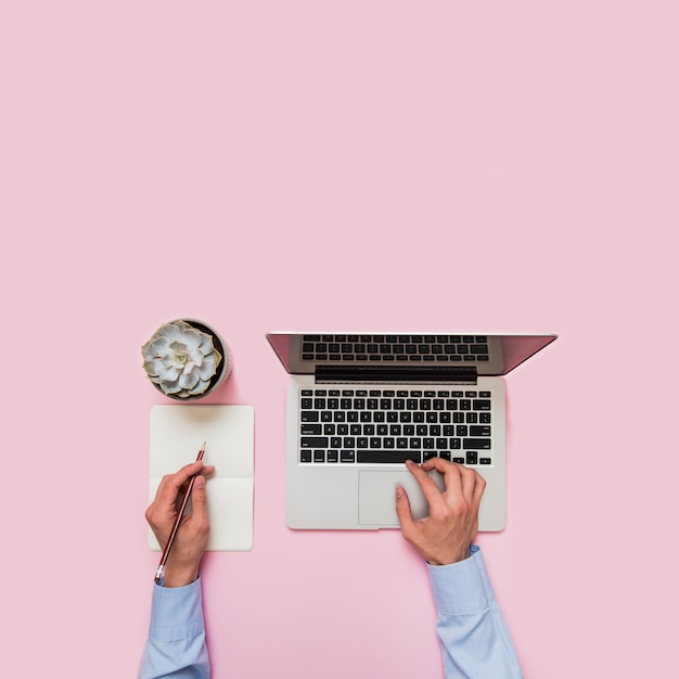 Close-up van de hand van een bedrijfspersoon het typen op laptop en het schrijven op papier met potlood tegen roze achtergrond