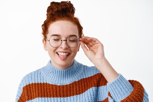 Close up van dom en schattig vrouwelijk model met rood haar gekamd in broodje en bril brillen frame aan te raken en tong met gesloten ogen staande op witte achtergrond te tonen