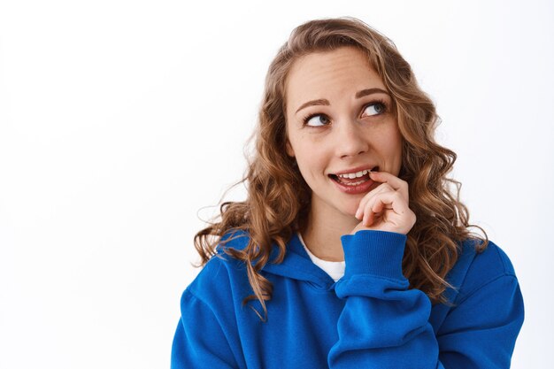 Close up van dromerige en flirterige vrouw bijtende vinger, kijkend naar de rechterbovenhoek nadenkend, een keuze maken, iets beslissen, over een witte muur staan