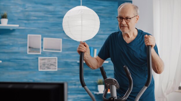 Close-up van een bejaarde man die thuis fietsoefeningen doet op een statische fiets. Senior persoon training met hometrainer voor wellness en uithoudingsvermogen. Gepensioneerde volwassene die fietsende cardio-machine gebruikt