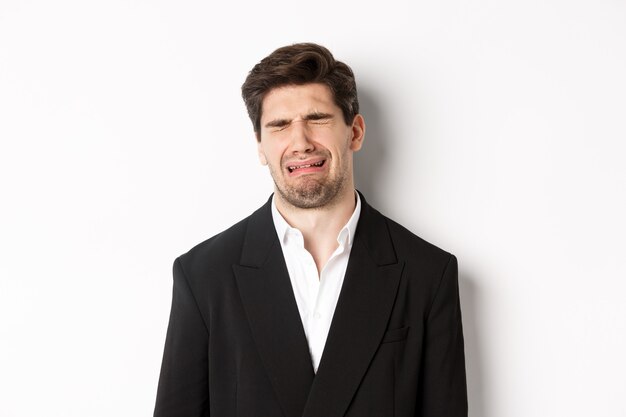Close-up van een ellendige man in pak, huilend en snikkend, verdrietig, staande tegen een witte achtergrond.