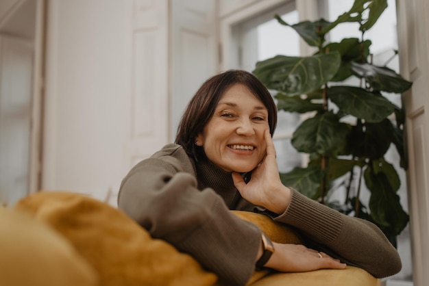 Gratis foto close-up van een gelukkige blanke vrouw van middelbare leeftijd die thuis op een comfortabele gele bank zit genieten van een positieve houding van kleine dingen