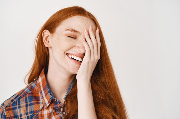 Close-up van een gelukkige jonge vrouw met rood natuurlijk haar en een bleke huid, vrolijk glimlachend en de helft van het gezicht bedekt, staande over een witte muur