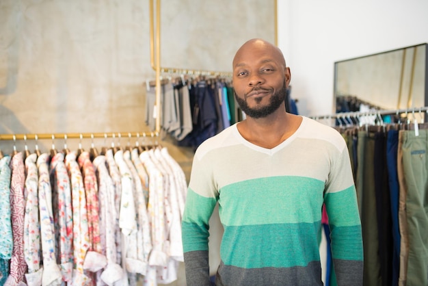 Close-up van een gelukkige man die vrije tijd doorbrengt in de kledingwinkel. knappe afrikaanse man wachten op consultant poseren voor camera in de buurt van hangers met shirts. kledingzaken, mannenmode en winkelconcept