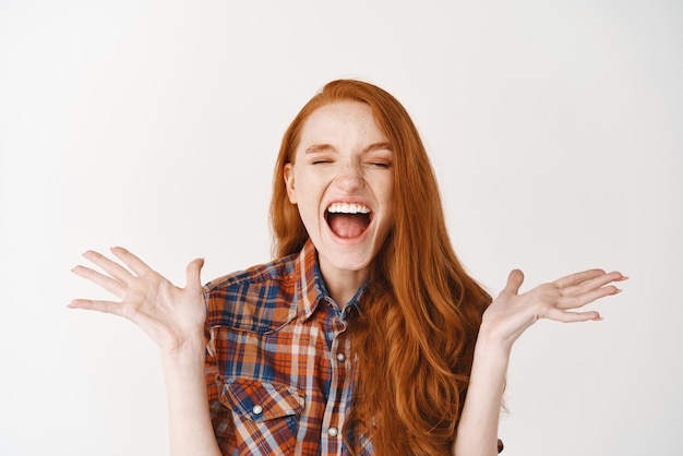 Close-up van een gelukkige roodharige vrouw die schreeuwt van vreugde en geluk en er verbaasd uitziet terwijl ze over een witte achtergrond staat
