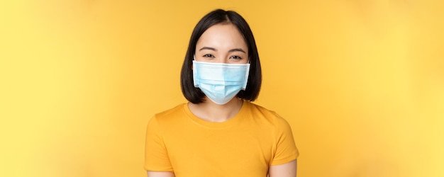 Close-up van een glimlachende gelukkige aziatische vrouw die een medisch gezichtsmasker draagt van covid in gele tshirt