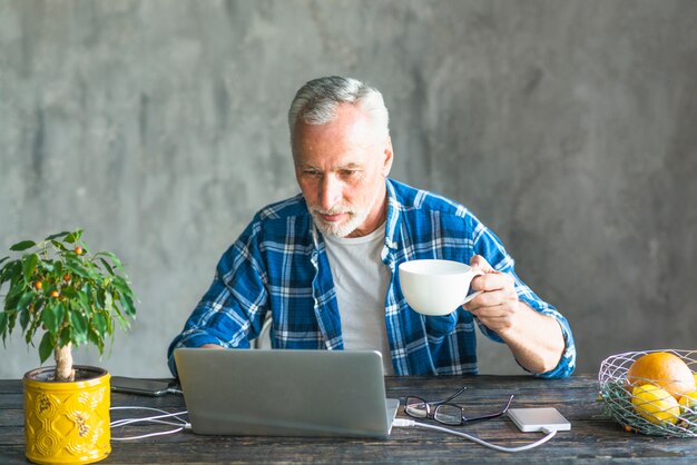 Close-up van een hogere man met koffiekopje met behulp van laptop belast met power bank