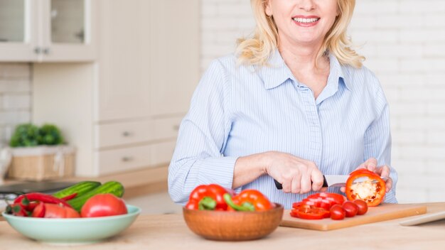 Close-up van een hogere vrouw die de rode groene paprika met mes op lijst in de keuken snijdt