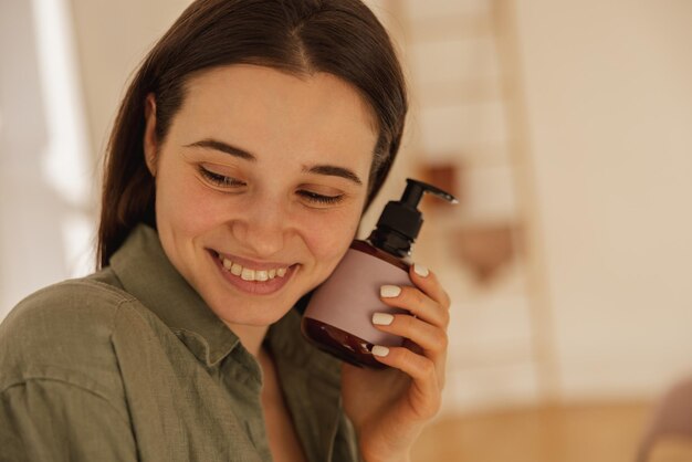 Close-up van een jong kaukasisch brunette meisje dat bescheiden glimlacht terwijl ze binnenshuis naar beneden kijkt Model in shirt met bodylotion Huidverzorging en hydratatie concept