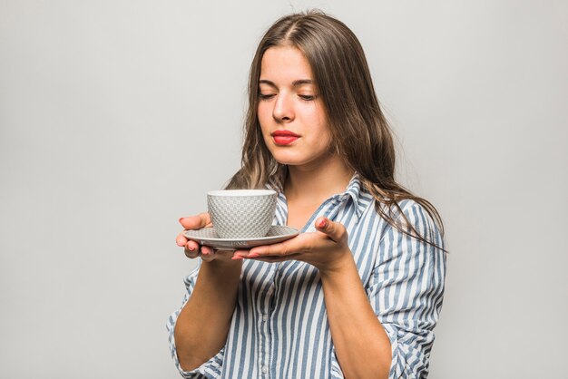 Close-up van een jonge vrouw die de kop thee en de schotel houdt tegen grijze achtergrond