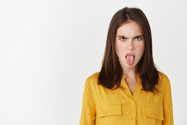 Close-up van een jonge vrouw die tong en teleurstelling toont, overstuur fronst, naar iets walgelijks staart, een afkeer uitdrukt terwijl ze over een witte muur staat.