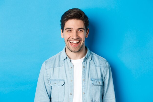 Close-up van een knappe jongeman die lacht, casual kleding draagt, over een blauwe achtergrond staat